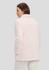 Chaqueta de forro polar rosa con una superficie texturizada y esponjosa, que cuenta con un cuello y mangas largas, llevada sobre pantalones de pijama con estampado.