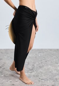 Crna maksi suknja sa naborima i visokim prorezom, s uvijenim detaljem sprijeda. Glatki materijal i prilagođeni kroj naglašavaju njezinu siluetu.
