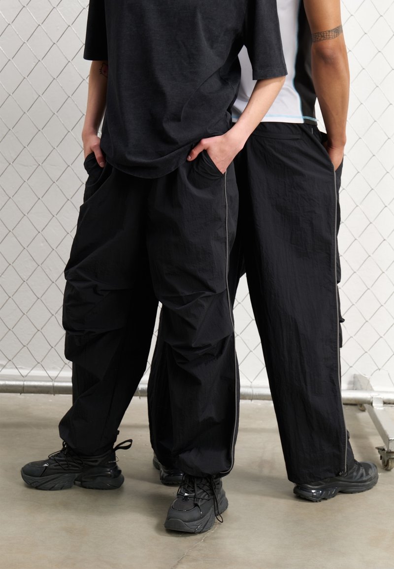 Svarta cargobyxor med en lös passform, texturerat tyg, sidofickor och kontrasterande vita sömmar, ihop med svarta sneakers.