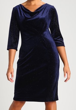 Navy blue velvet knee-length dress with three-quarter sleeves and a draped neckline, worn by a standing woman against a plain background.