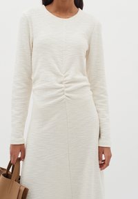 Långärmad klänning i texturerat vitt tyg, med en central samlad detalj vid midjan. Modellen håller en liten brun handväska.