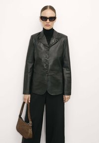 Blazer in pelle nera con colletto a patta, chiusura con sei bottoni e maniche lunghe, abbinato a pantaloni neri a gamba larga e una borsa marrone con texture.