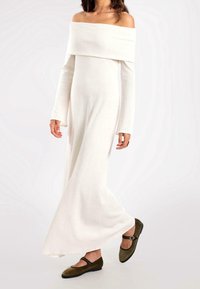 Robe blanche à manches longues et décolletée, fabriquée en tissu côtelé, avec un corsage ajusté et une jupe fluide. Des chaussures vert foncé viennent compléter le look.