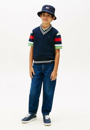 Jongen met een navy blauwe bucket hat, een mouwloos navy blauw vest over een gestreept shirt met korte mouwen, losse blauwe jeans en navy blauwe sneakers staat rustig.