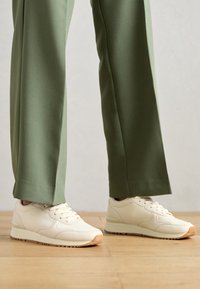 Vita sneakers med en slät läderfinish, rund tå och gummisula. I kombination med gröna byxor med vida ben. Bakgrund av trägolv.