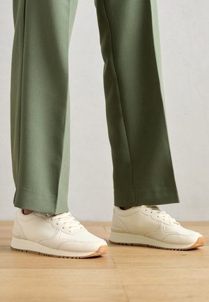 Baskets blanches avec une finition en cuir lisse, bout rond et semelle en caoutchouc. Assorti à un pantalon vert à large jambe. Fond de parquet en bois.