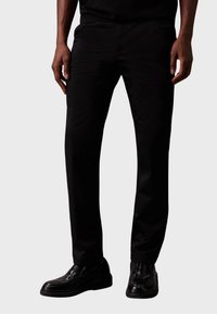 Zwarte broek van gladde stof, met een rechte pasvorm en zakken. Gecombineerd met glanzende zwart leren schoenen.