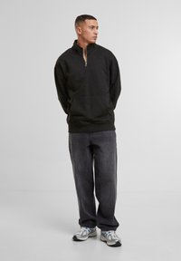 Black zip-neck sweatshirt with long sleeves and front pocket, paired with loose dark gray jeans and silver running shoes with blue accents.