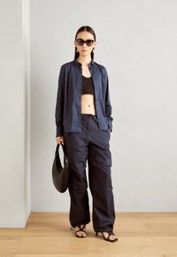 Camicia blu con bottoni sopra un crop top nero, abbinata a pantaloni blu larghi, borsa nera e sandali neri con lacci su un pavimento di legno.