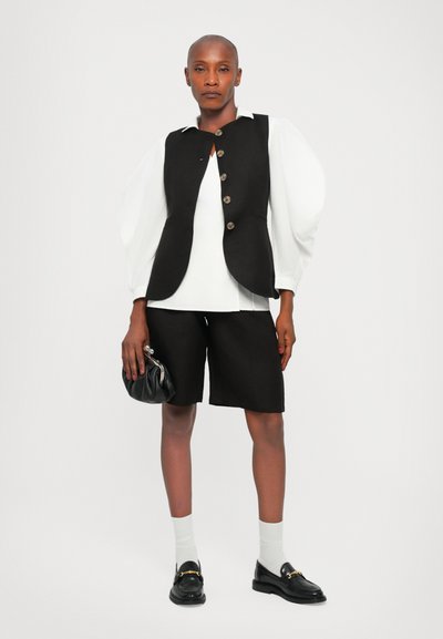 Svart vest med knapper over en hvit topp, kombinert med svarte shorts, svarte loafers og en svart håndveske. Glatt stoff og strukturert design.