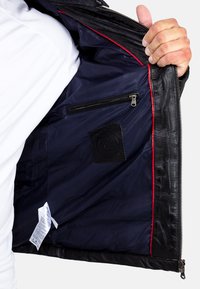 Interno di una giacca di pelle nera, con fodera blu navy, cuciture rosse, tasca con zip e un'etichetta di stoffa nera con logo.