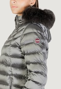 Kobieta nosząca błyszczącą srebrną kurtkę typu puffer z kapturem wykończonym futrem oraz czerwoną naszywką z logo "COLMAR" na górnej części rękawa.
