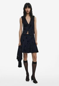 Ensemble deux pièces marine comprenant un veston sans manches ajusté avec un décolleté en V profond et une jupe évasée à motifs floraux. Assorti avec des bas jusqu'aux genoux.