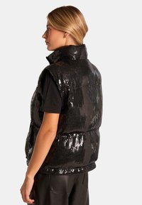 Schwarze Paillettenweste mit hohem Kragen, gepolstertem Design und sichtbaren Stichen. Getragen über einem schwarzen Hemd, weist sie eine glänzende, strukturierte Oberfläche auf.