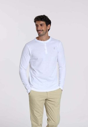 Chemise henley blanche à manches longues avec un col boutonné, associée à un pantalon beige clair. Texture douce en coton, minimalisme dans le branding.