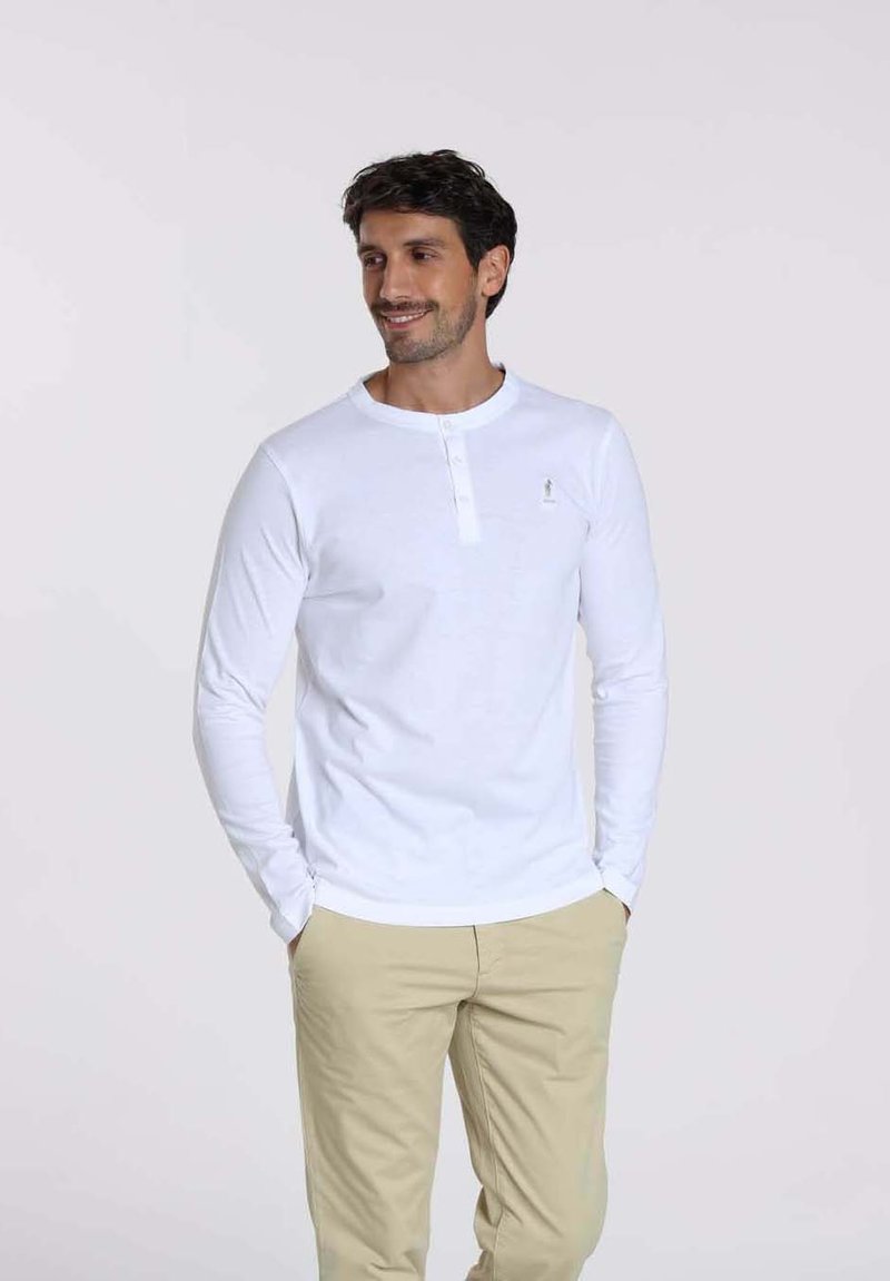Chemise henley blanche à manches longues avec un col boutonné, associée à un pantalon beige clair. Texture douce en coton, minimalisme dans le branding.