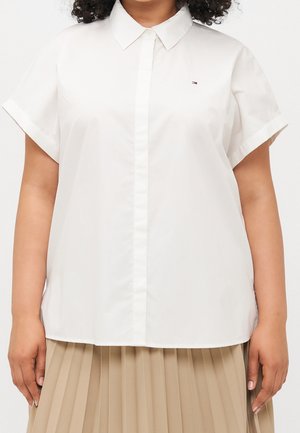 Person wearing a short-sleeve white button-up shirt with a small logo on the chest and a beige pleated skirt.