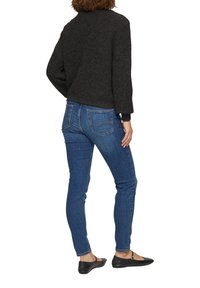 Dunkles Strickpullover, eng anliegende blaue Jeans, flache schwarze Schuhe. Die Textur ist weich; die Jeans haben Gesäßtaschen und ein lässiges Design.