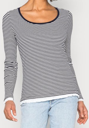 Mujer con una camiseta de rayas horizontales blancas y negras de manga larga con cuello redondo y vaqueros azul claro.