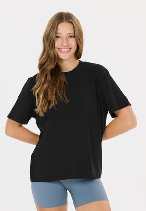 Smilende ung kvinde med langt bølget hår iført en løs sort t-shirt og tætsiddende blå shorts mod en ensfarvet hvid baggrund.