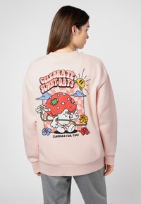Rosa Sweatshirt mit einem bunten Grafikdesign auf der Rückseite, das einen lächelnden Pilz, Blumen und den Text "Feiere sonnige Tage" zeigt.