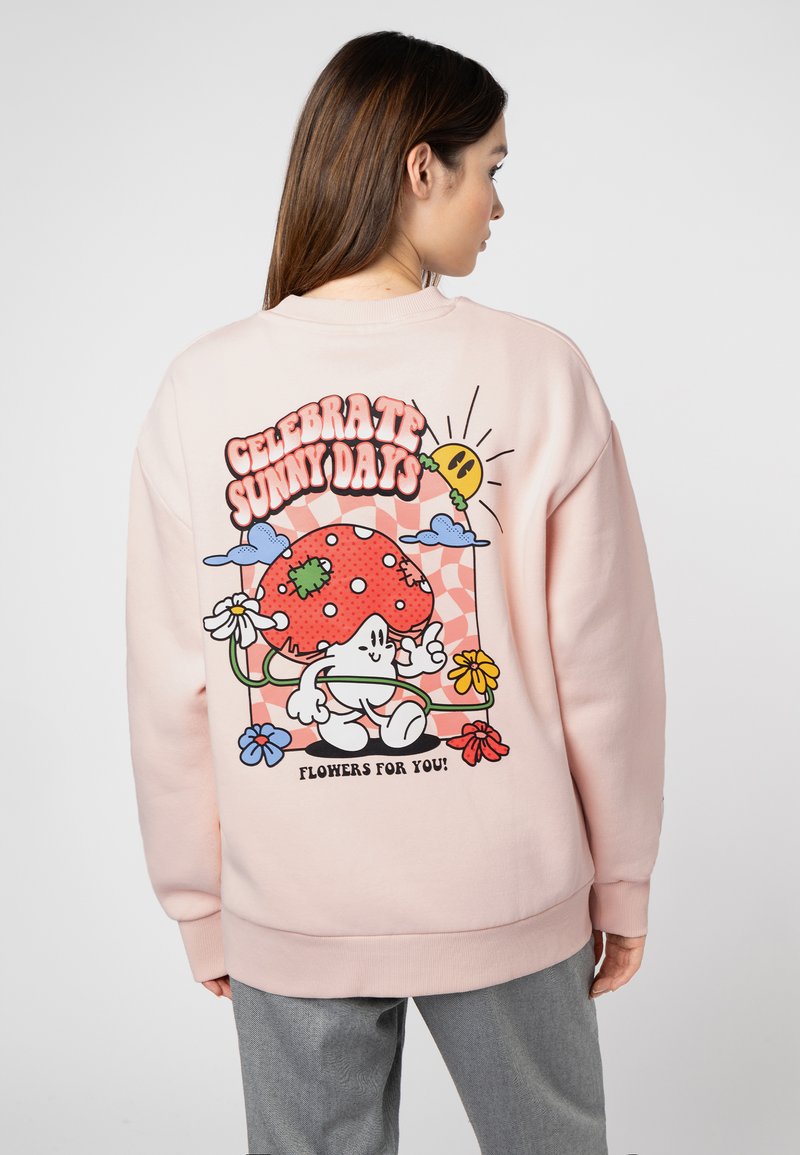 Rosa Sweatshirt mit einem bunten Grafikdesign auf der Rückseite, das einen lächelnden Pilz, Blumen und den Text "Feiere sonnige Tage" zeigt.