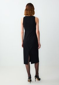 Schwarzes ärmelloses Kleid mit taillierter Silhouette, knielang, mit einem Schlitz im Rücken. Getragen mit schwarzen High Heels und transparenten Strumpfhosen.