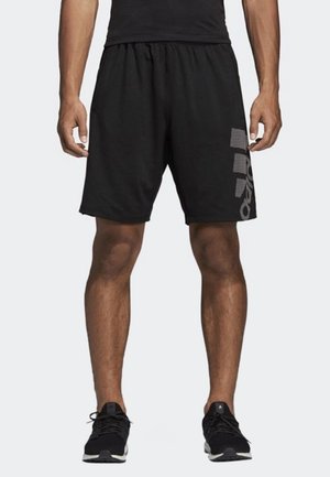 Mann trägt schwarze sportliche Shorts mit einem grauen Muster am linken Bein, schwarze Turnschuhe, steht vor einem neutralen hellen Hintergrund.