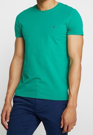 Mężczyzna w dopasowanym zielonym t-shircie z krótkim rękawem z małym logo na piersi oraz w ciemnoniebieskich spodniach, stojący na jasnym, jednolitym tle.