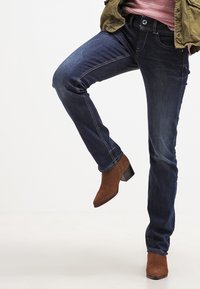 Jeans de denim en azul oscuro con un corte recto, que presentan costuras en contraste y bolsillos delanteros. Llevados con botas de ante marrón hasta el tobillo.