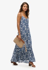 Robe maxi à motif bleu et blanc avec des bretelles spaghetti, un ourlet volanté et un tissu léger. Accessoirisée avec une pochette tissée et des sandales marron.