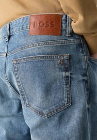 Jeans de mezclilla azul claro con una cinturilla de cuero marrón grabada con "BOSS" y un pequeño acento de logotipo metálico cerca del bolsillo trasero.