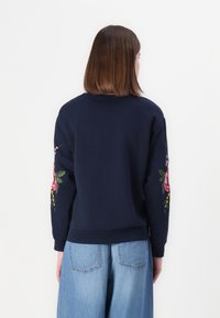 Persona con cabello castaño lacio lleva un sudadera azul marino con bordados florales rosas en las mangas y unos jeans de pierna ancha de color azul claro, de espaldas.