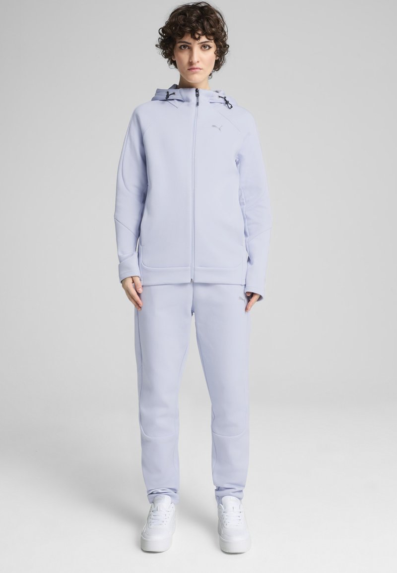 Veste de sport lilas clair avec capuche, fermeture éclair à l'avant, et deux poches latérales, assortie à un pantalon fuselé et des baskets blanches.