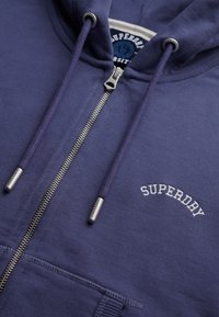 Felpa blu con zip e texture spazzolata. Presenta una zip argentata, un cordino e il logo "SUPERDRY" ricamato in bianco sul petto.