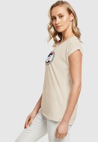 Beige bomulds t-shirt med et rundt grafisk design, der viser et farverigt portræt omgivet af en blomsterkant. Korte ærmer og afslappet pasform.