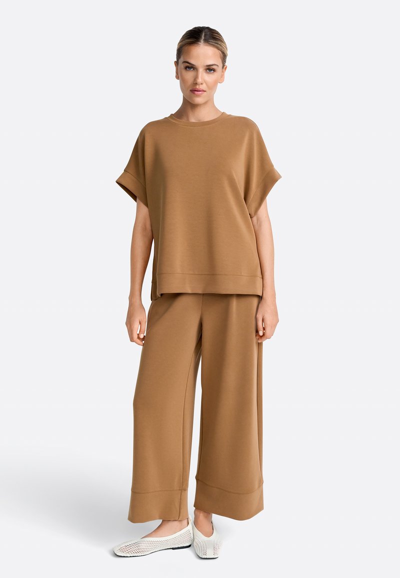 Mujer de pie con camiseta holgada marrón de manga corta, pantalones de pierna ancha y zapatos blancos de malla tipo slip-on, sobre un fondo claro y liso.