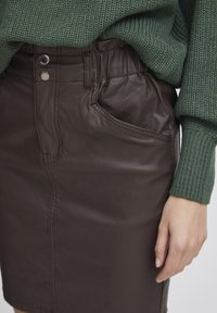 Jupe en faux cuir marron avec poche latérale, taille haute et deux fermetures à boutons argentés. Pull vert à côtes partiellement visible au-dessus.