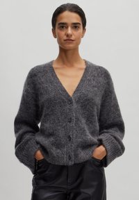 Grijze gebreide cardigan met een V-hals, lange mouwen en vijf zwarte knopen. Zachte textuur en relaxed fit, gedragen over hoge zwarte broeken.