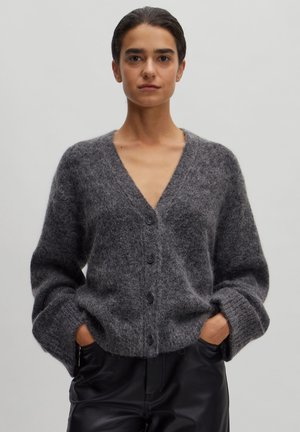 Grauer, gestrickter Cardigan mit V-Ausschnitt, langen Ärmeln und fünf schwarzen Knöpfen. Weiche Textur und lockere Passform, getragen über hochtaillierten schwarzen Hosen.