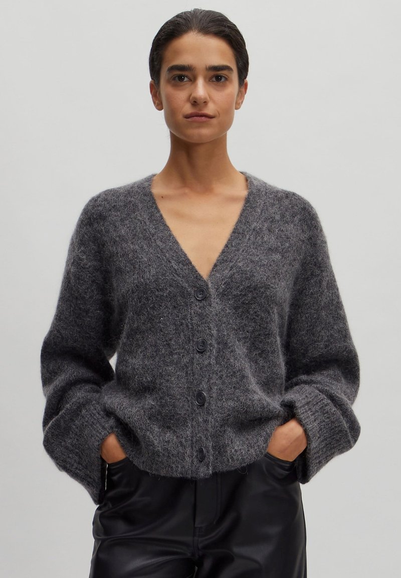 Grijze gebreide cardigan met een V-hals, lange mouwen en vijf zwarte knopen. Zachte textuur en relaxed fit, gedragen over hoge zwarte broeken.