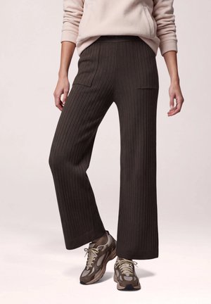 Le mannequin porte un pantalon côtelé marron foncé à jambes larges, un sweat-shirt clair et des baskets marron, devant un fond de studio uni.