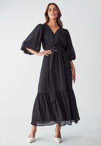 Black pleated maxi dress with a V-neck, balloon sleeves, a belted waist featuring a circular buckle, and a tiered hem. Smooth, lightweight fabric.