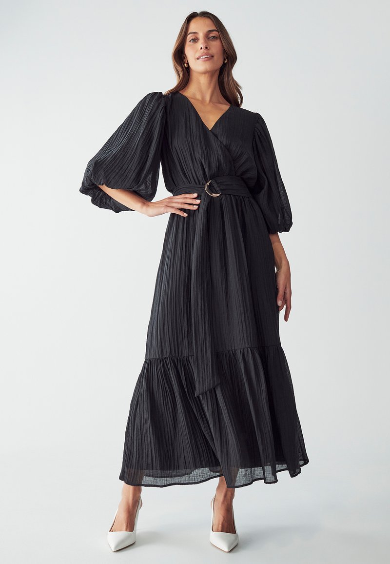 Robe maxi plissée noire avec un décolleté en V, des manches ballon, une taille ceinturée avec une boucle circulaire, et un ourlet à volants. Tissu lisse et léger.