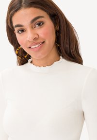 Top bianco a coste con collo alto e orlo arricciato, dotato di maniche lunghe. Orecchini grandi a cerchio in tartaruga completano l'outfit.
