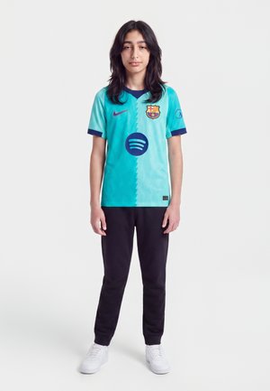 Jeune personne debout, portant un maillot de football turquoise et bleu de Barcelone, un pantalon noir et des baskets blanches sur fond blanc.