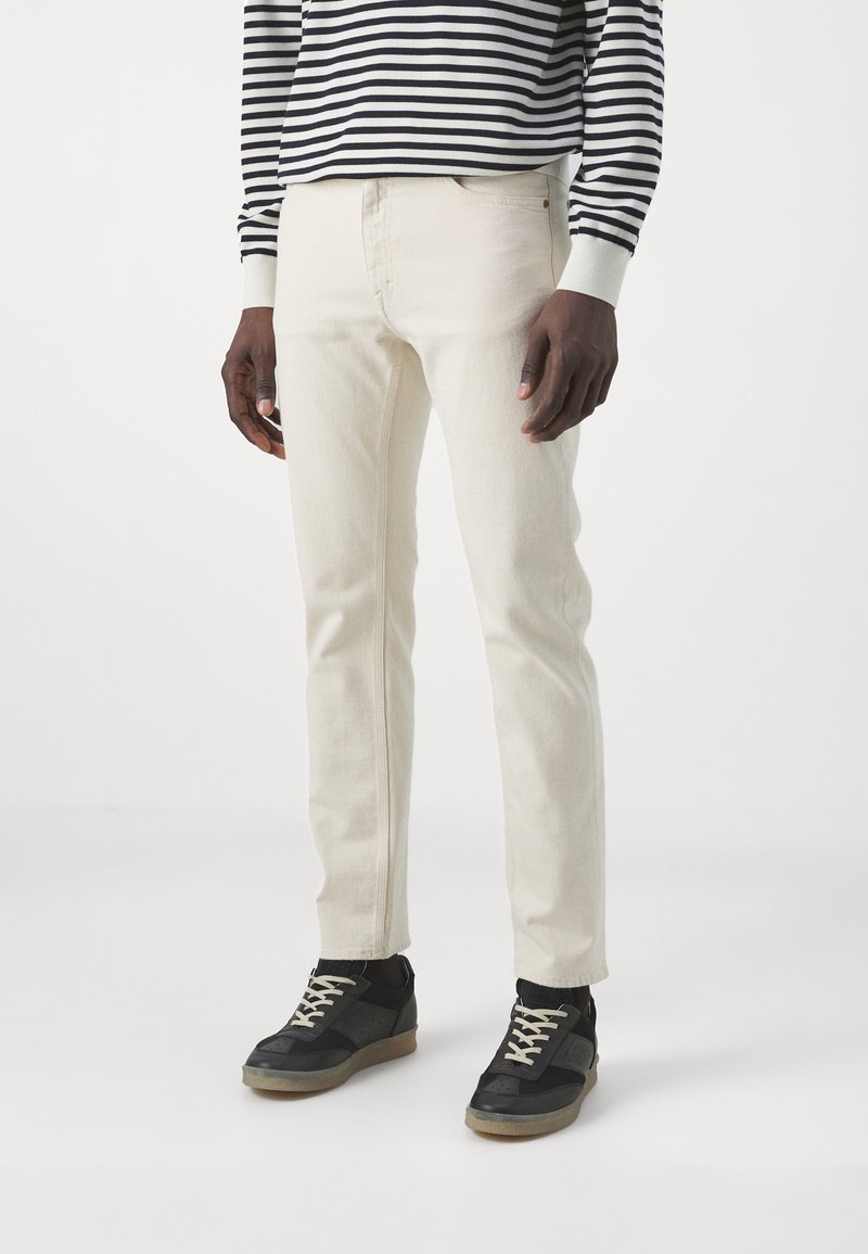 Jeans color beige claro con un corte slim, con una textura suave y costuras mínimas, combinados con zapatillas negras con detalles grises.