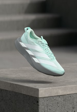 Világoskék futócipő fehér csíkokkal és szürke talppal, amely egy betonlépcső felett lebeg, háttérben elmosódott lépcsőkkel.