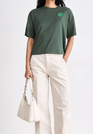 Groen kort t-shirt met een ronde halslijn en een rond geborduurd embleem, gecombineerd met crème hoge taillebroeken en een witte handtas.
