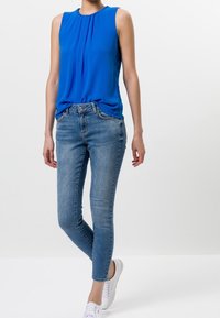 Top azul sin mangas con pliegues, combinado con jeans ajustados azul claro y zapatillas blancas. Tela suave, ajuste casual y diseño sencillo.